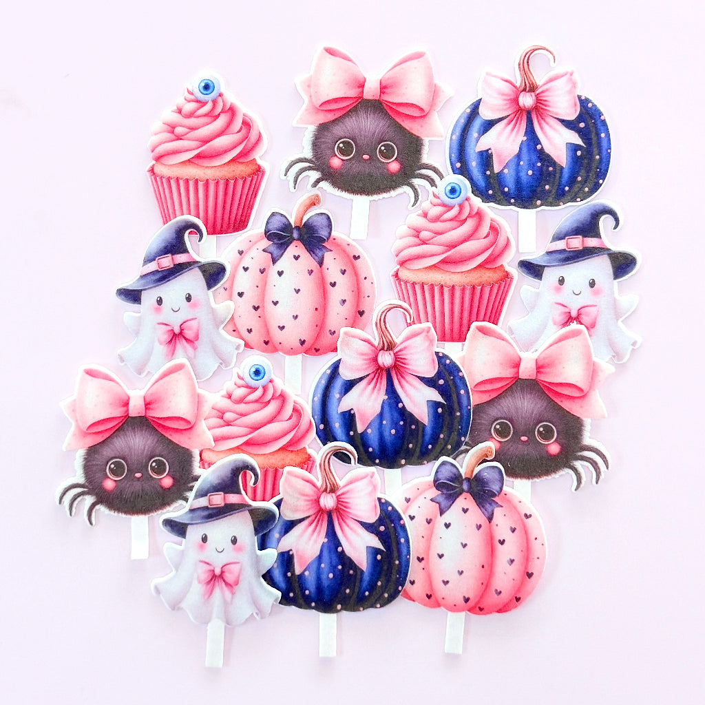 A set of cute Halloween-themed paper cupcake toppers on sticks, featuring pink cupcakes, dark blue pumpkins with pink bows, black spiders, and white ghosts with pink and purple hats, arranged on a light pink background. Edible Wafer Card Cupcake Toppers - Cute Pink Halloween